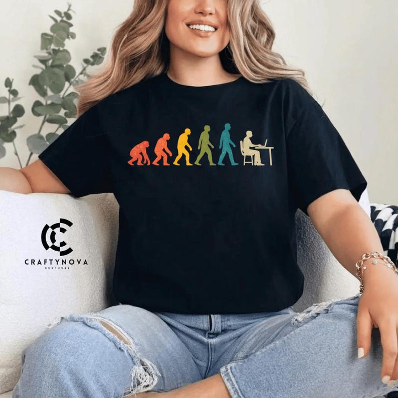 Peut inclure: Un t-shirt noir &agrave; manches courtes avec une impression graphique color&eacute;e de l'&eacute;volution humaine. Le motif montre la progression d'un primate vers un humain assis &agrave; un bureau avec un ordinateur, en silhouettes orange, jaune, verte et bleue.