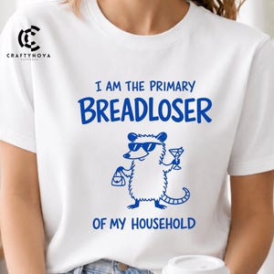 Könnte beinhalten: Ein weißes T-Shirt mit kurzen Ärmeln und einer blauen Grafik eines Opossums mit Sonnenbrille, das eine Handtasche und ein Martiniglas hält. Der Text lautet I AM THE PRIMARY BREADLOSER OF MY HOUSEHOLD in blauen Buchstaben. Ein Logo ist oben links.