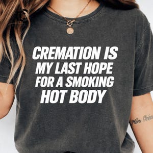 Op de afbeelding: Een antracietgrijs T-shirt met korte mouwen en de witte tekst "CREMATION IS MY LAST HOPE FOR A SMOKING HOT BODY" in een vet, schreefloos lettertype op de borst. Het kledingstuk heeft een ronde hals en een gewassen, vintage uitstraling.