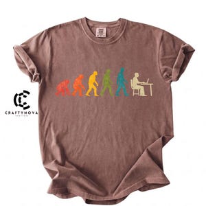 Peut inclure: Un t-shirt marron &agrave; manches courtes arborant un graphique color&eacute; de l'&eacute;volution humaine. Le motif montre une progression allant d'un primate &agrave; un humain assis &agrave; un bureau avec un ordinateur portable, en silhouettes textur&eacute;es rouge, orange, jaune, vert et bleu.
