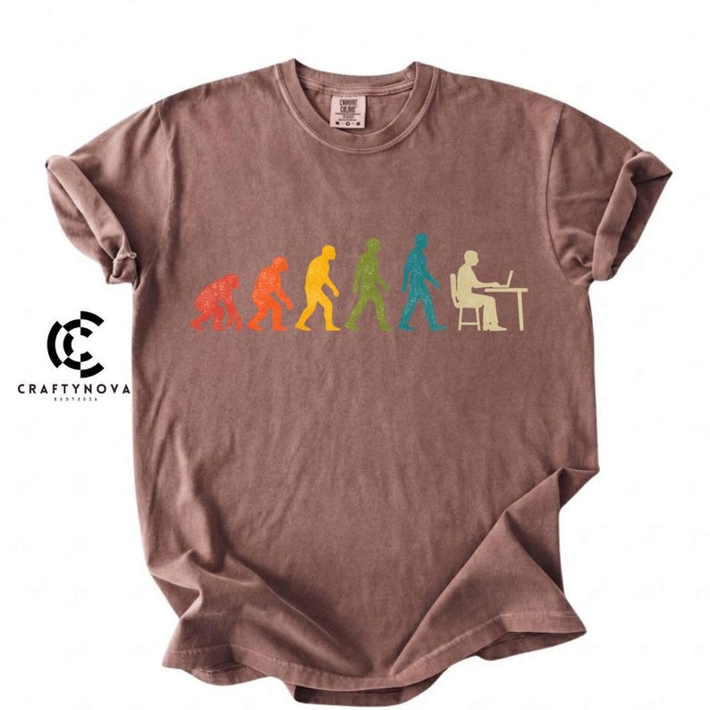 Peut inclure: Un t-shirt marron &agrave; manches courtes arborant un graphique color&eacute; de l'&eacute;volution humaine. Le motif montre une progression allant d'un primate &agrave; un humain assis &agrave; un bureau avec un ordinateur portable, en silhouettes textur&eacute;es rouge, orange, jaune, vert et bleu.