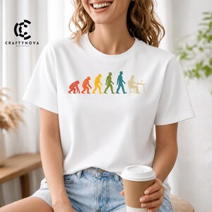 Peut inclure: Un t-shirt blanc &agrave; manches courtes avec une impression graphique color&eacute;e de l'&eacute;volution humaine, allant d'un primate &agrave; une personne assise &agrave; un bureau avec un ordinateur. Le motif pr&eacute;sente une texture vieillie et un spectre de couleurs incluant le rouge, l'orange, le jaune, le vert et le bleu.