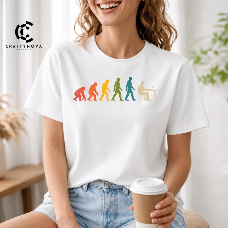 Peut inclure: Un t-shirt blanc &agrave; manches courtes avec une impression graphique color&eacute;e de l'&eacute;volution humaine, allant d'un primate &agrave; une personne assise &agrave; un bureau avec un ordinateur. Le motif pr&eacute;sente une texture vieillie et un spectre de couleurs incluant le rouge, l'orange, le jaune, le vert et le bleu.