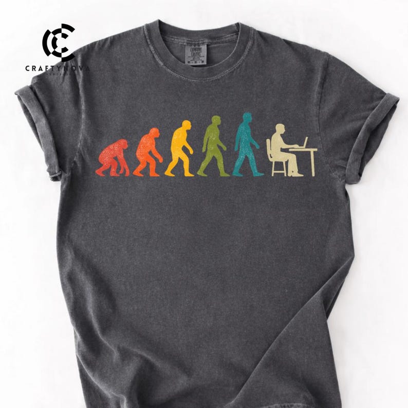 Peut inclure: Un t-shirt gris anthracite arborant un graphique color&eacute; de l'&eacute;volution humaine, commen&ccedil;ant par un primate rouge et progressant &agrave; travers des silhouettes orange, jaune, verte et bleue, finissant par une silhouette blanche assise &agrave; un bureau.