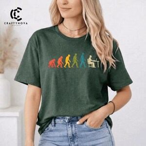 Peut inclure: Un t-shirt &agrave; manches courtes vert fonc&eacute; avec une impression graphique repr&eacute;sentant l'&eacute;volution humaine, partant d'un primate pour aboutir &agrave; une personne assise &agrave; un bureau avec un ordinateur. Le motif utilise des tons orange, jaune, vert et bleu.