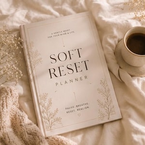 Könnte beinhalten: Ein beigefarbener Soft Reset Planner mit floralen Akzenten und dem Text "Soft Reset Planner". Eine Tasse Kaffee auf einer Untertasse befindet sich neben dem Planer. Auf dem Cover steht "A Gentle Reset For Your Mind & Life".