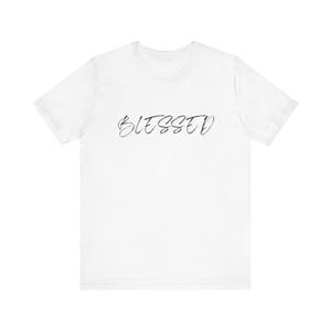 May include: A white short-sleeved t-shirt featuring the word BLESSED printed in a black, thin, cursive script font across the centre of the chest. The garment has a standard crew neckline and a plain, solid white background fabric design.