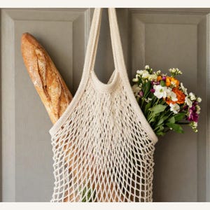 May include: A beige, open-weave shopping bag filled with a fresh baguette and a colorful bouquet of flowers. The bag hangs on a gray door, suggesting a trip to the market or a delivery.