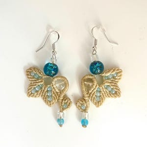 May include: A pair of dangle earrings featuring beige macrame cord knotted into a leaf-like shape, accented with light blue seed beads, a clear glass bead, and a round, speckled turquoise glass bead, all suspended from silver-toned metal hooks.