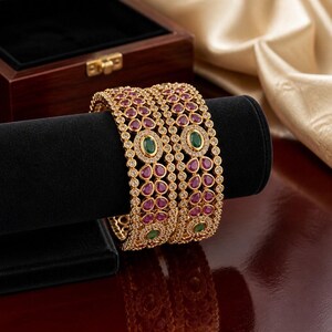 May include: Two gold-tone bangles featuring a repeating pattern of teardrop-shaped pink stones and circular green stones. The metal framework is intricately detailed with small clear stones set along the edges and between the larger colored gems.