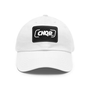 Pode incluir: Um boné de beisebol branco com um emblema retangular preto no painel frontal. O emblema exibe o texto branco CNQR. em uma fonte sem serifa em negrito, contido dentro de um contorno retangular branco com cantos arredondados.