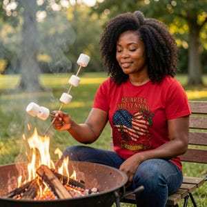 May include: A woman wearing a red t-shirt with a patriotic design, roasting marshmallows over a campfire. The shirt features an American flag graphic and the text "Quarter Millennial EST. 1776". She is sitting on a wooden bench.