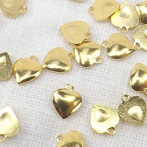 May include: A group of 20 gold heart-shaped charms with a loop at the top. The charms are scattered on a white fabric background.