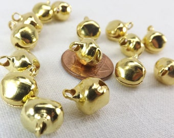 Op de afbeelding: Een groep van 12 gouden belletjes met een lus aan de bovenkant. De belletjes zijn verspreid over een wit oppervlak, met een Amerikaanse penny voor de schaal.