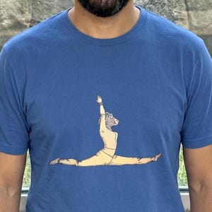 Könnte beinhalten: Ein blaues T-Shirt mit Rundhalsausschnitt und einer grafischen Illustration eines Primaten mit menschlichem Körper, der eine Yoga-Spagat-Pose ausführt. Die Figur trägt ein hellgelbes Outfit und hat einen Arm zum Himmel gestreckt.