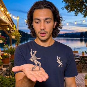 Könnte beinhalten: Ein dunkelblaues Kurzarm-T-Shirt mit einem Grafikdruck einer Giraffe, die ein gestreiftes Oberteil und eine Hose trägt und in einer Tanzpose auf der Brust sowie in einer kleineren Version auf der linken Brustseite abgebildet ist.