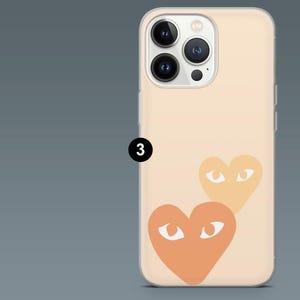 May include: A peach-colored phone case featuring two orange hearts with white eye designs. The case has a clear border and covers the back and sides of the phone. The phone's camera lenses are visible.