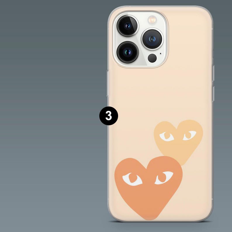 May include: A peach-colored phone case featuring two orange hearts with white eye designs. The case has a clear border and covers the back and sides of the phone. The phone's camera lenses are visible.