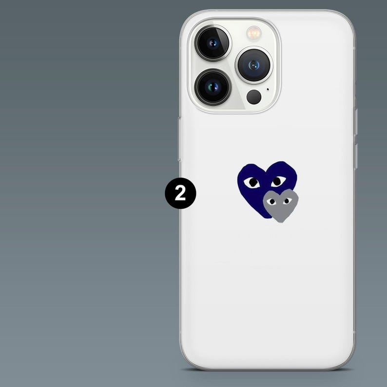 May include: White iPhone case with a navy blue heart design and a smaller gray heart. The case has a clear, protective design and features the phone's camera and flash. The design is simple and modern.