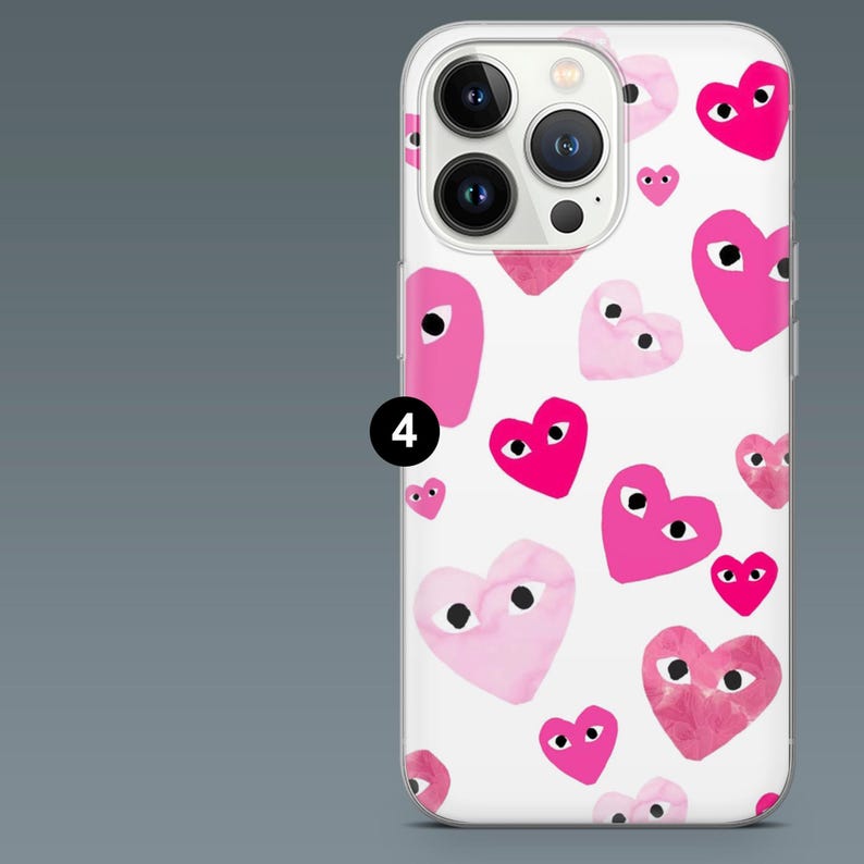May include: White iPhone case with a repeating pattern of pink hearts with black eyes. The hearts vary in size and shade, creating a playful design. The case has a clear border and a camera cutout.