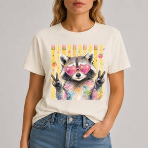 May include: A cream-colored t-shirt featuring a graphic of a raccoon wearing pink heart-shaped sunglasses. The raccoon is making a peace sign with both paws. The background of the graphic includes yellow vertical stripes, small stars, and apples.