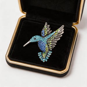 May include: A hummingbird brooch featuring a silver-toned metal frame encrusted with small, vibrant blue, turquoise, and lime green rhinestones. The bird is depicted in flight with wings extended, resting inside a square black jewelry box with gold trim.