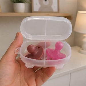 May include: A clear plastic pacifier case containing two pacifiers. One pacifier is a deep rose color, and the other is bright pink. The case is oval-shaped with a divided interior.