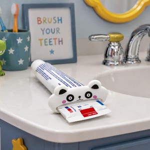 May include: A white panda-shaped toothpaste tube squeezer with a tube of toothpaste. The bathroom counter also features a blue toothbrush holder with toothbrushes, a green dinosaur, and a framed sign that says "BRUSH YOUR TEETH".