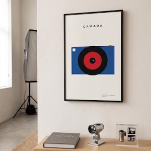 Puede incluir: Una lámina enmarcada con un gráfico minimalista de una cámara azul con un objetivo grande rojo y negro. El texto CAMARA aparece en la parte superior, con texto más pequeño en la parte inferior. La lámina tiene un marco negro en una pared blanca.