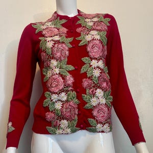 Puede incluir: Un cárdigan rojo con mangas largas. El suéter presenta diseños florales bordados en tonos rosa, verde y blanco. El bordado floral se concentra en la parte delantera y las mangas. El cárdigan tiene un cierre de botones.