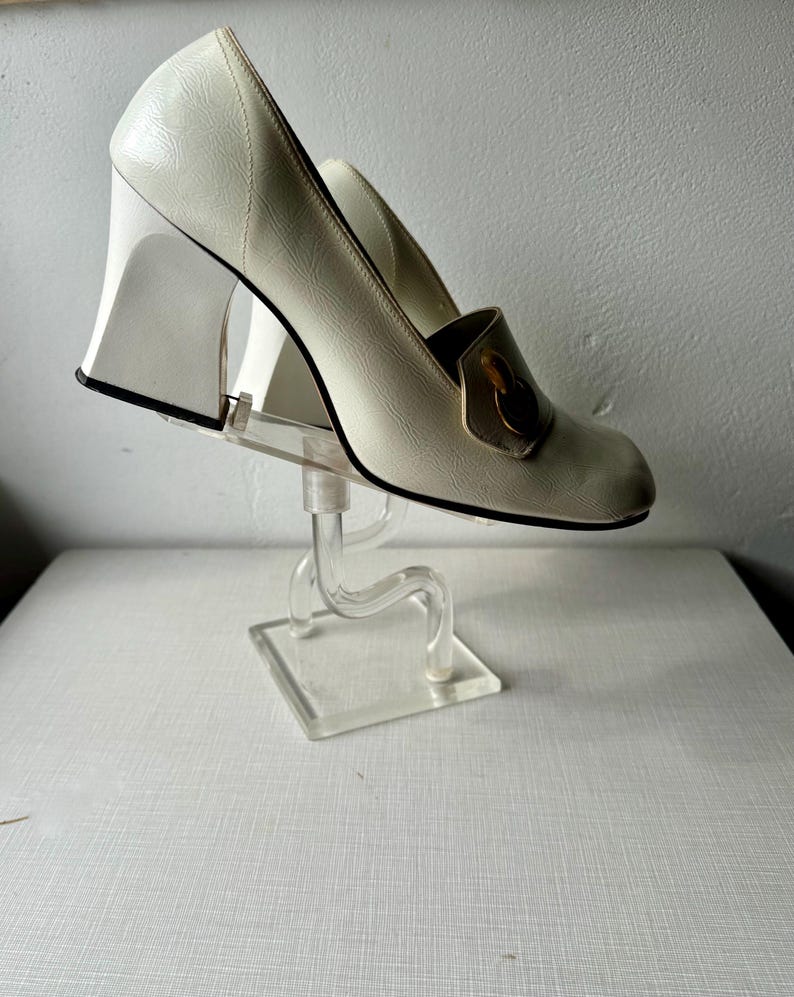 K&ouml;nnte beinhalten: Wei&szlig;e Lederschuhe mit Absatz und goldener Schnalle. Die Schuhe stehen auf einem durchsichtigen Acryl-Schuhst&auml;nder.