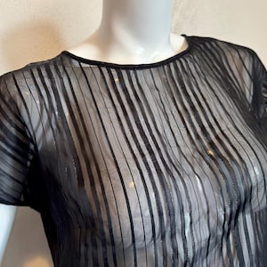 Può includere: Top nero trasparente a righe verticali. La blusa a maniche corte ha uno scollo tondo ed è realizzata in un tessuto leggero e trasparente. Il top è esposto su un manichino.