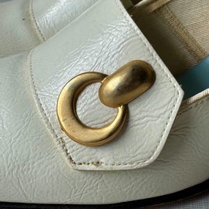 K&ouml;nnte beinhalten: Wei&szlig;e Lederschuhe mit einer goldenen Metallschnalle in Ringform. Die Schuhe haben eine spitze Zehenpartie und einen niedrigen Absatz.