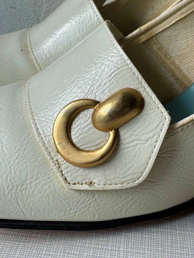 K&ouml;nnte beinhalten: Wei&szlig;e Lederschuhe mit einer goldenen Metallschnalle in Ringform. Die Schuhe haben eine spitze Zehenpartie und einen niedrigen Absatz.