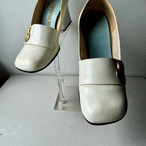 K&ouml;nnte beinhalten: Ein Paar wei&szlig;e Lederschuhe mit goldenen Schnallen. Die Schuhe haben einen dicken Absatz und eine quadratische Spitze. Der Markenname "Tom Maids" ist auf der Innenseite eines Schuhs sichtbar.