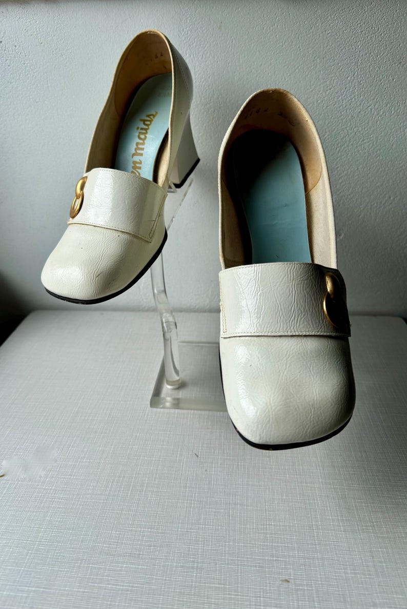 K&ouml;nnte beinhalten: Ein Paar wei&szlig;e Lederschuhe mit goldenen Schnallen. Die Schuhe haben einen dicken Absatz und eine quadratische Spitze. Der Markenname "Tom Maids" ist auf der Innenseite eines Schuhs sichtbar.