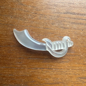 May include: A clear, translucent, decorative object shaped like a small sword or saber. The handle has a detailed, ridged design, and the blade curves gracefully. The object is lying on a wooden surface.