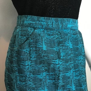 May include: A teal blue skirt with a geometric pattern. The skirt has two pockets on the front.