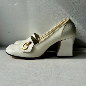 K&ouml;nnte beinhalten: Wei&szlig;e Lederschuhe mit Absatz und einem goldenen Metallring-Detail. Die Schuhe haben einen klobigeren Absatz.