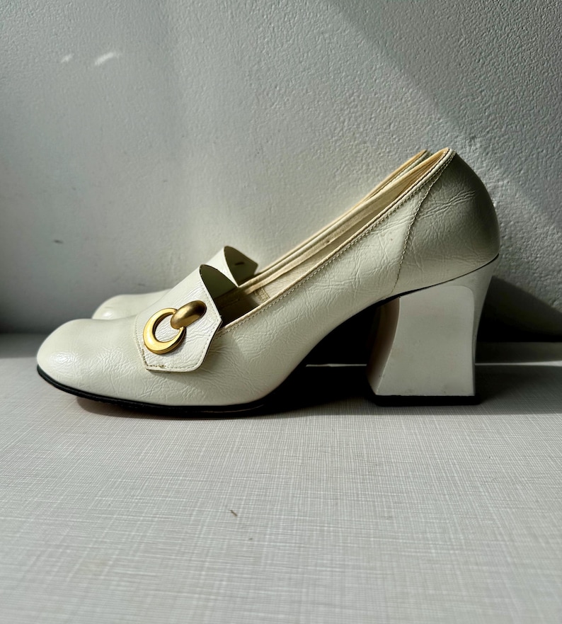 K&ouml;nnte beinhalten: Wei&szlig;e Lederschuhe mit Absatz und einem goldenen Metallring-Detail. Die Schuhe haben einen klobigeren Absatz.