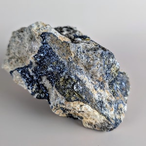May include: A rough, irregularly shaped rock with a mix of colors. The rock features areas of dark blue, gray, and tan, with a slightly reflective, metallic sheen in some spots. The overall texture appears coarse and natural.