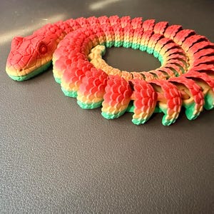 May include: A 3D-printed articulated snake toy in vibrant colors. The snake's head is red with yellow and green accents, and its body transitions from red to yellow and green. The segmented design allows for flexible movement.