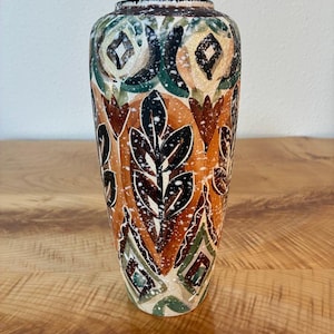 May include: A ceramic vase featuring a tapered cylindrical shape with a hand-painted pattern of dark brown leaves, geometric diamond shapes, and floral motifs in shades of orange, green, and cream, finished with a white speckled paint splatter.