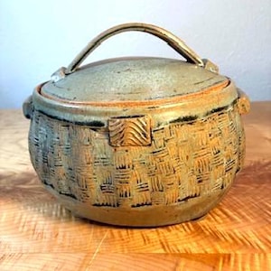 May include: A round ceramic lidded jar with a textured basket-weave pattern on the exterior. The vessel features a muted green and brown glaze, two small side handles, and a curved handle on the lid. The surface has a matte, earthy finish.
