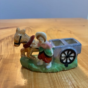 Puede incluir: Una figura de cerámica que muestra un burro color canela tirando de un carro azul con una persona que lleva un gorro blanco, una blusa rosa y una falda roja. El burro tiene un arnés oscuro y el carro tiene una rueda negra sobre una base verde.