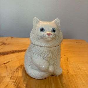 May include: A white ceramic cat-shaped jar with a removable lid. The cat has long, sculpted fur texture, bright blue eyes, a small pink nose, and whiskers. The figure is depicted in a seated position with its tail curled around its body.