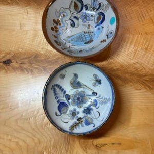 May include: Two round ceramic bowls featuring hand-painted blue and brown floral and bird motifs. The top bowl has a brown rim and a light blue bird, while the bottom bowl displays a central bird surrounded by blue flowers and a dark blue rim.