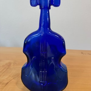 May include: A cobalt blue glass bottle shaped like a violin or cello. The bottle features a long neck, a rounded body with moulded details representing strings and f-holes, and a top opening that mimics the scroll and pegbox of a string instrument.