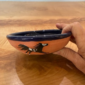 May include: A small, round, terracotta-colored ceramic bowl with a dark blue glazed rim. The exterior features a hand-painted black horse with white spots in a running pose. The interior of the bowl is also finished in a dark blue glaze.