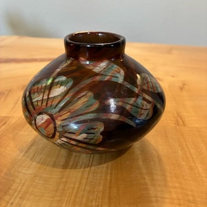 Pode incluir: Um pequeno vaso de cerâmica castanho-escuro, de forma redonda e acabamento brilhante. A superfície apresenta um padrão floral pintado em tons suaves de verde, azul e branco, com pinceladas visíveis. O vaso tem um pescoço curto e estreito.
